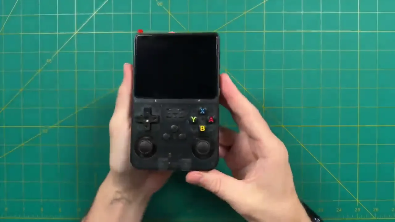 Console R36S noire posée sur un tapis de découpe, écran éteint, vue de dessus